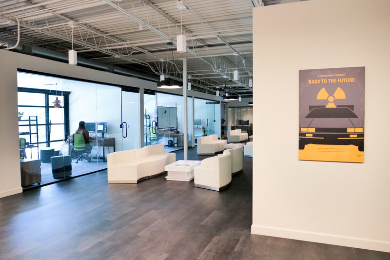 A clean hallway filled with white couches. Behind glass windows and doors,employees work on computers next to large windows that let in a lot of sunlight. On the hallway wall is a minimalistic Back To The Future film poster.