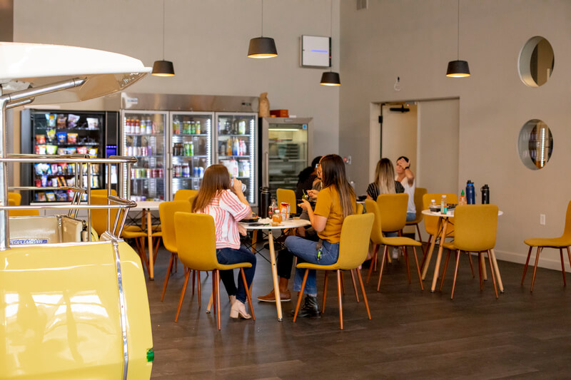 A group of seated employees happily socializes in the company eating area, filled with small tables and yellow chairs in front of a row of vending machines and well-stocked beverage coolers