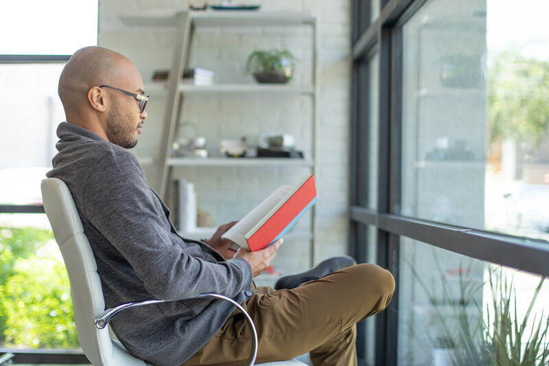 A man in a gray hoodie thoughtfully reads a red book while seated